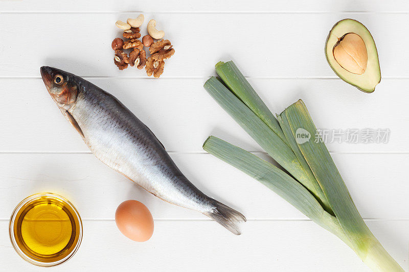 选择含有omega - 3和不饱和脂肪的食物来源。欧米茄3，欧米茄6。含有健康脂肪的产品:亚麻籽油，鳄梨，杏仁，腰果，开心果，鱼，鸡蛋，韭菜。俯视图，白色木板